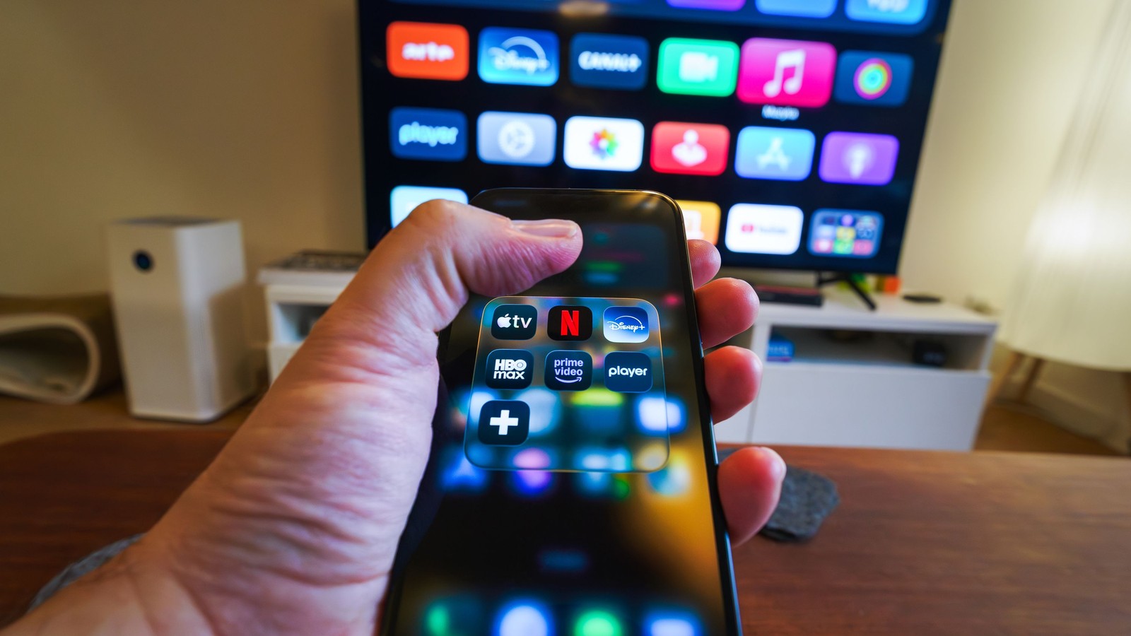 Tangan memegang telefon pintar yang memaparkan folder aplikasi penstriman di hadapan televisyen.